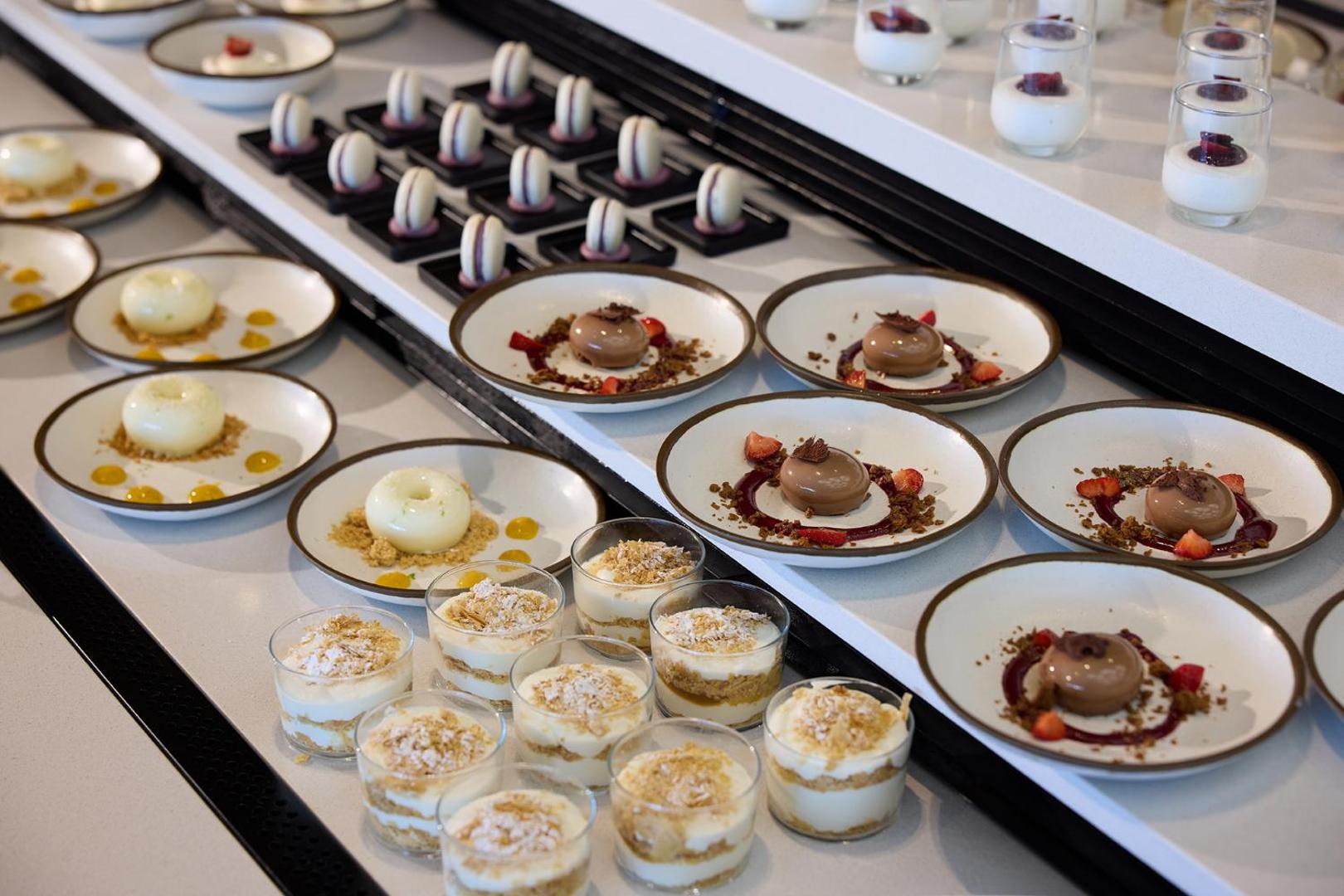 Krāšņa desertu bufete ar dažādām kūkām, uzpūteņiem un makarūniem uz balta galda.