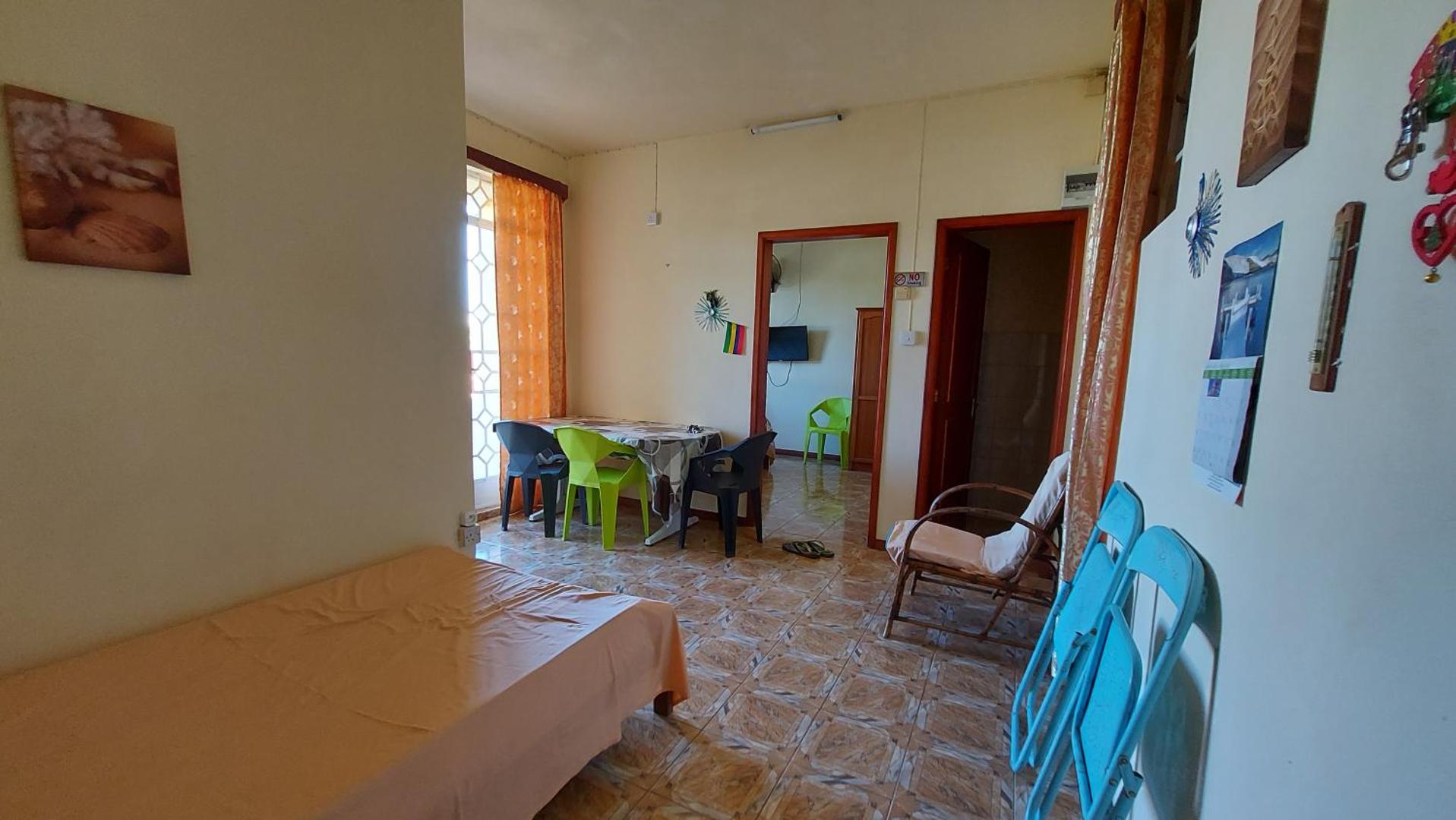 Dzīvojamā istaba ar gultu, ēdamgaldu un krāsainiem krēsliem flīžu grīdas telpā.