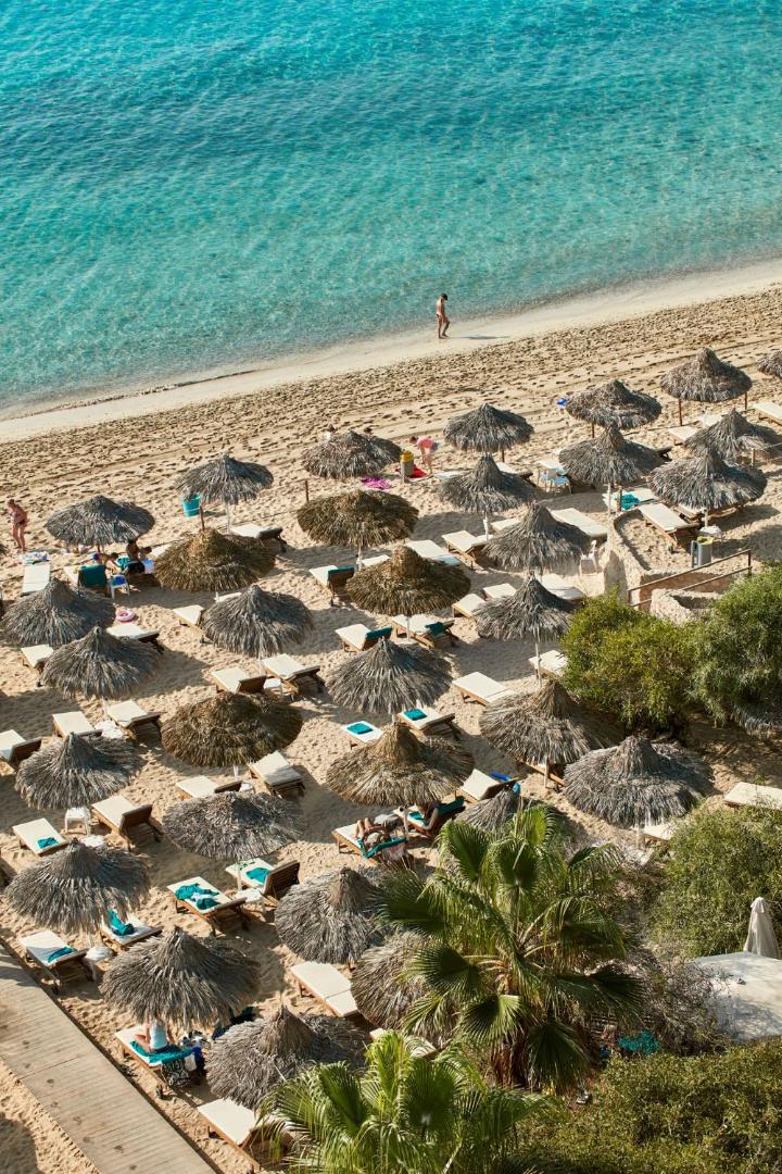 Skats no augšas uz Grecian Bay pludmali Kiprā — daudzi salmu saulessargi smiltīs blakus tirkīzzilam jūras ūdenim un zaļām palmām