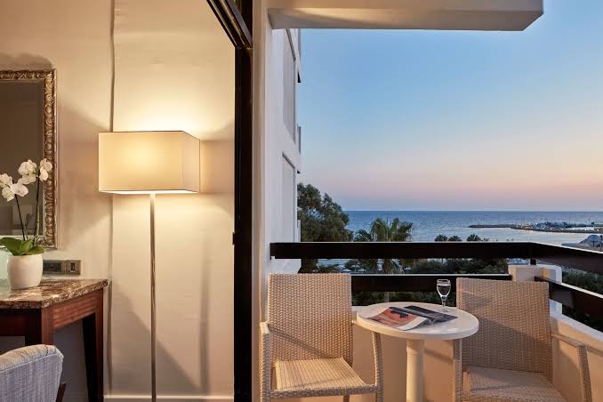 Grecian Bay viesnīcas balkons saulrieta laikā ar skatu uz jūru — divi pīti krēsli, galdiņš un mājīgs apgaismojums vakarā