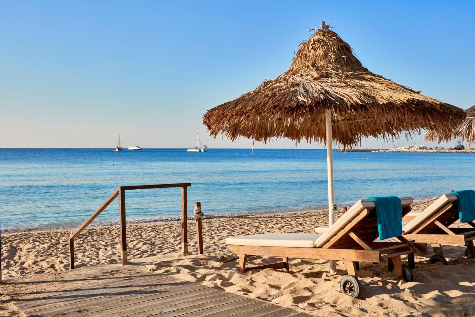 Grecian Bay viesnīcas privātā pludmale Ajanapā ar koka sauļošanās krēsliem, salmu saulessargiem un skatu uz tirkīzzilo Vidusjūru un jahtām