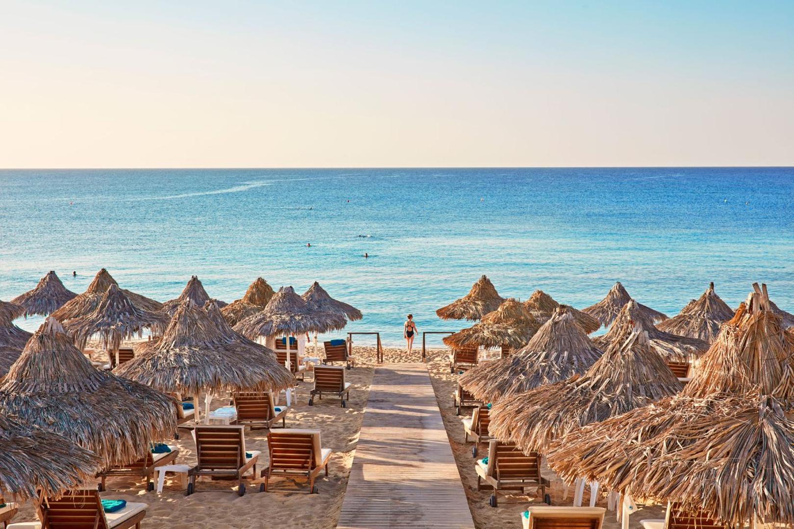 Grecian Bay viesnīcas privātā pludmale Aijanapā, Kiprā — koka laipa ved caur salmu saulessargiem un zviļņiem uz zilo Vidusjūru