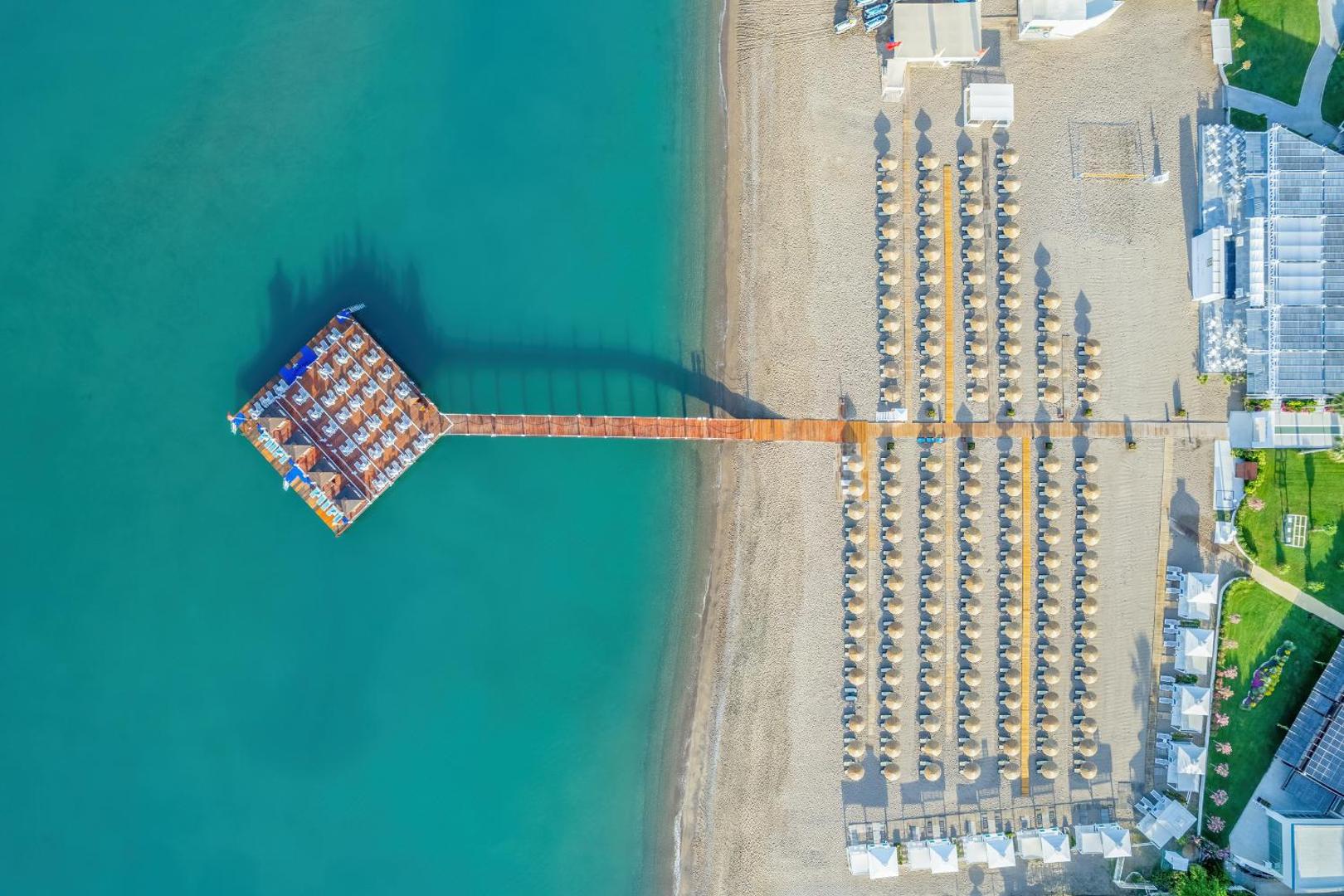 Aerofoto skats uz Baia Lara smilšu pludmali, saulessargiem un garu koka piestātni jūrā.