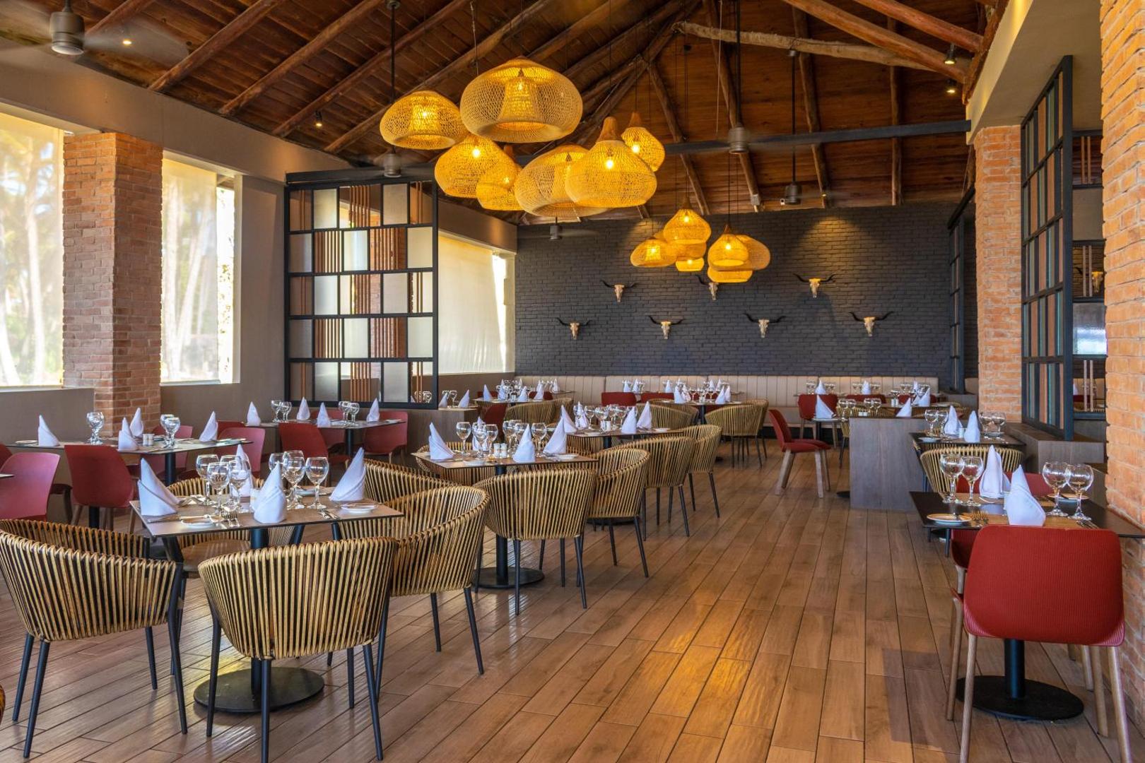 Restorāna interjers ar koka griestiem, pītiem gaismekļiem un eleganti klātiem galdiem.