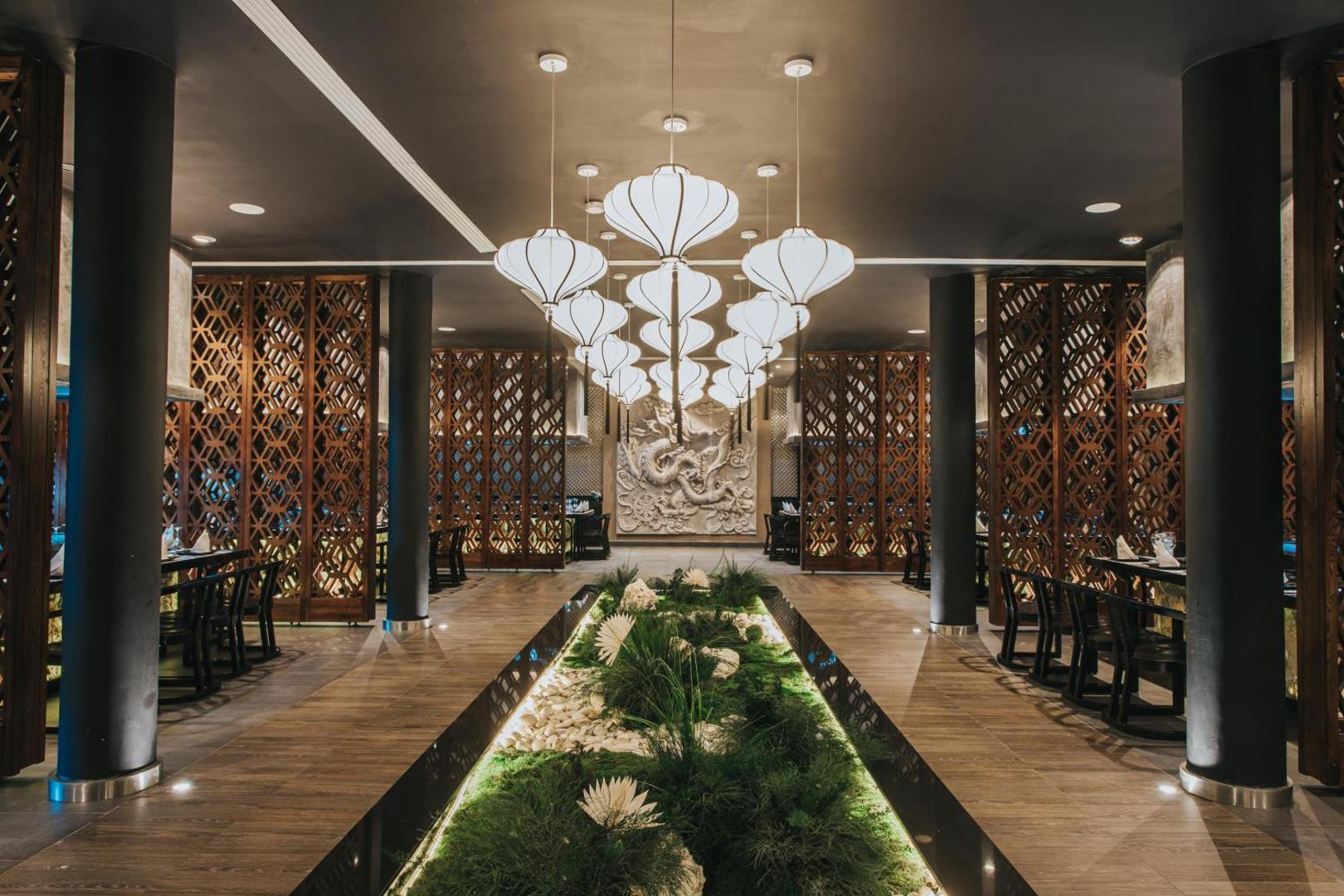 Eleganta restorāna ieeja ar baltām laternām, koka paneļiem un dekoratīvu augu joslu.