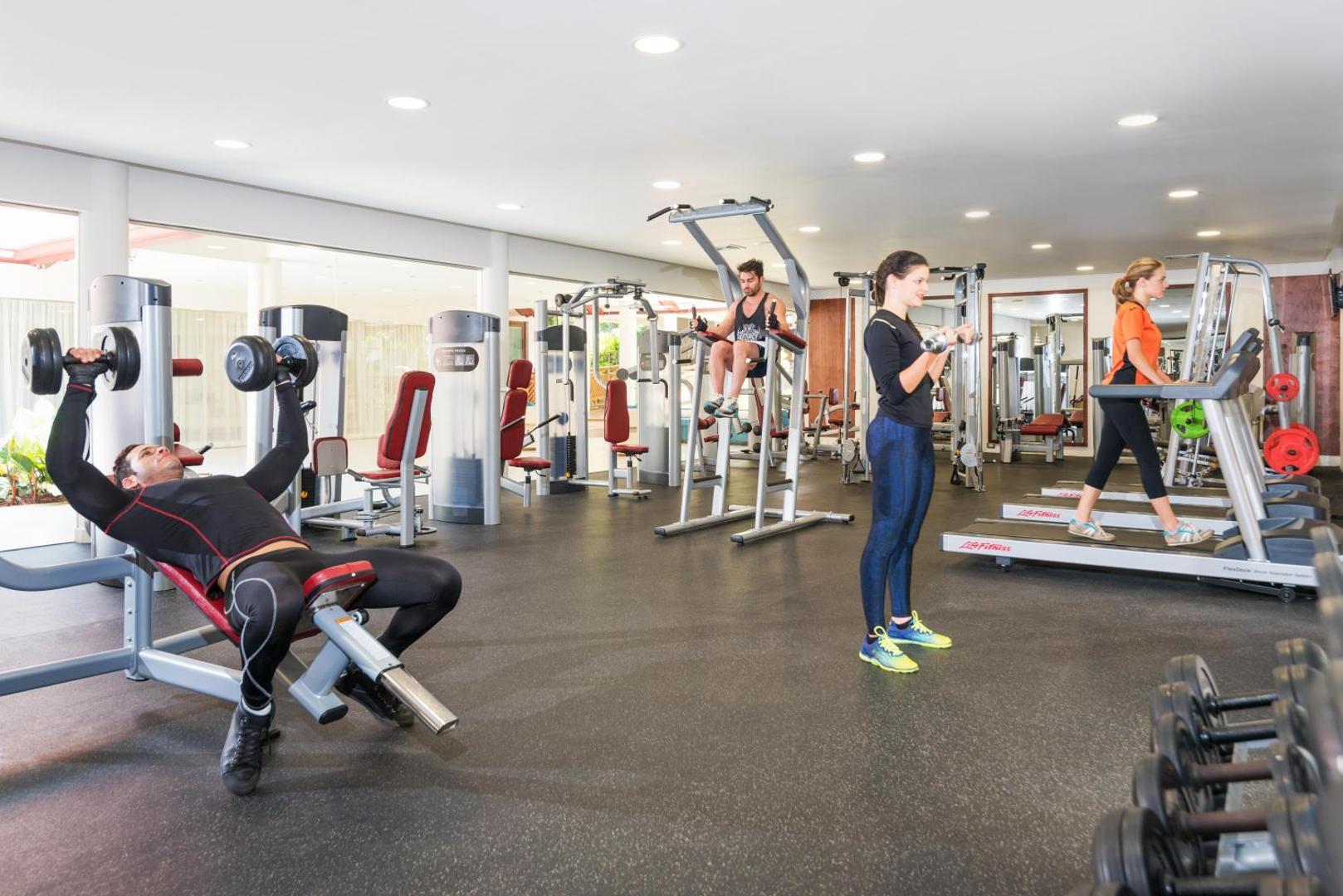 Mūsdienīga viesnīcas trenažieru zāle ar dažādiem fitnesa aparātiem un cilvēkiem, kas sporto.