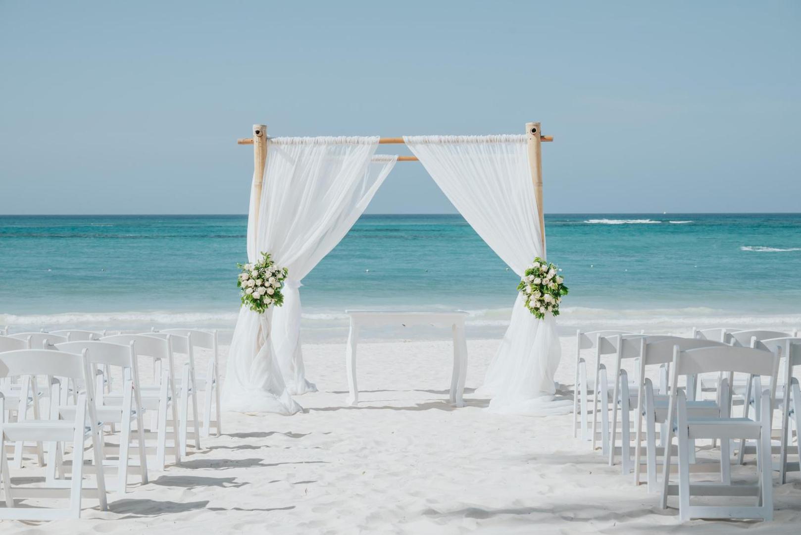 Balta kāzu arka ar ziediem un krēsliem smilšainā pludmalē pie tirkīzzila ūdens.