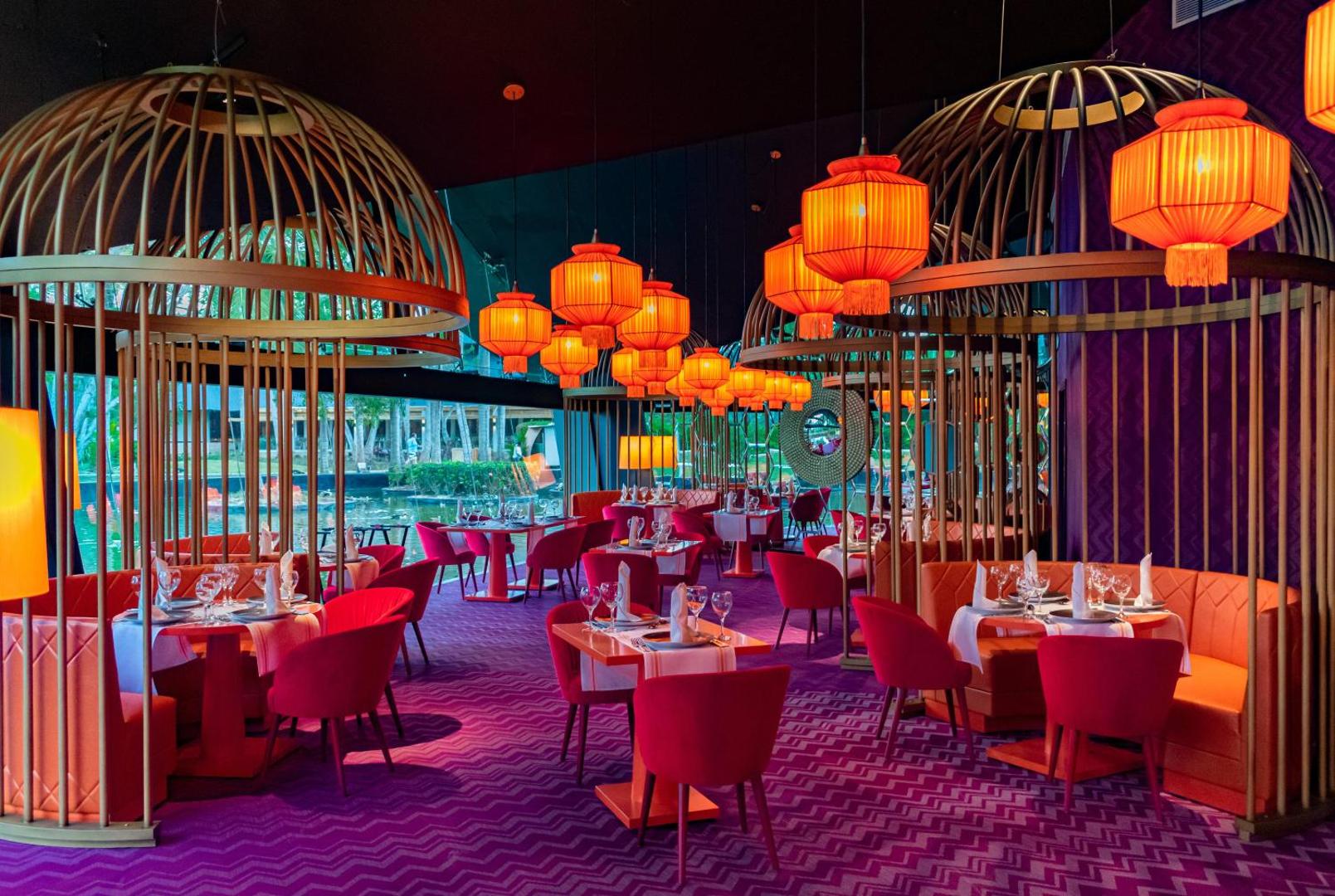 Tematisks restorāns ar purpursarkanu paklāju, sarkaniem krēsliem un lieliem dekoratīviem būriem.