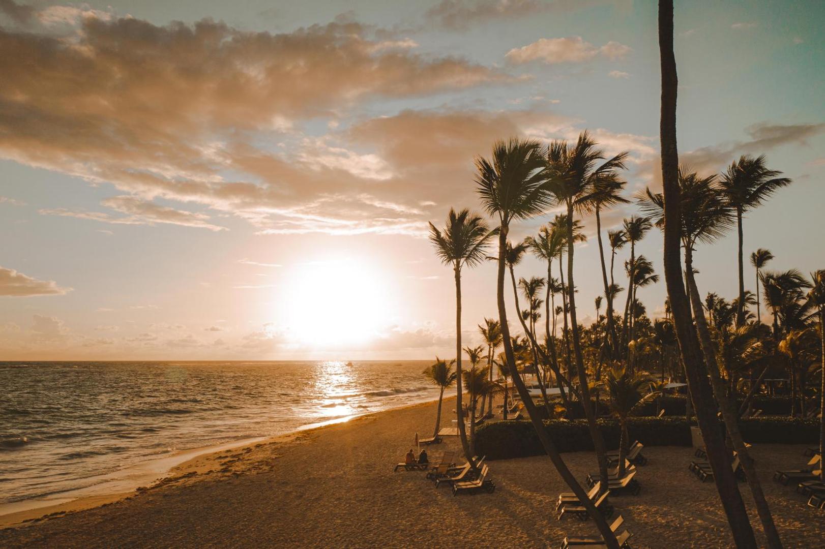 Saulriets smilšainā pludmalē ar palmām un sauļošanās krēsliem pie okeāna.