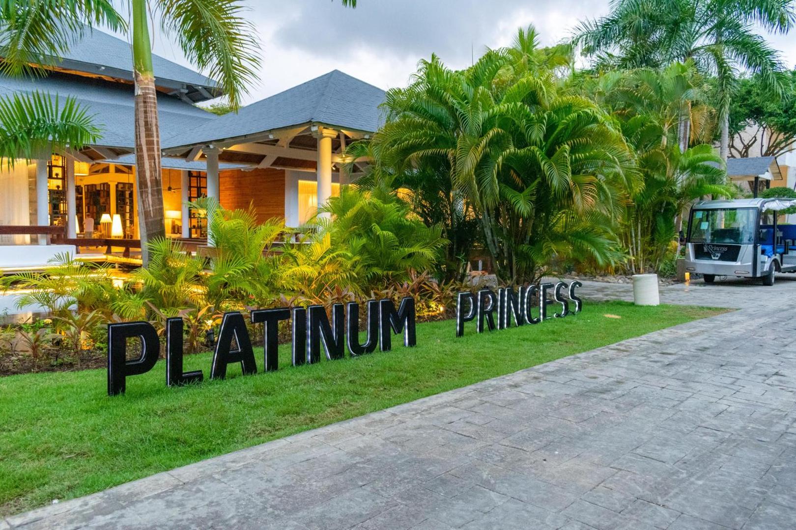 Platinum Princess uzraksts uz kopta zālāja pie viesnīcas ēkas un palmām.