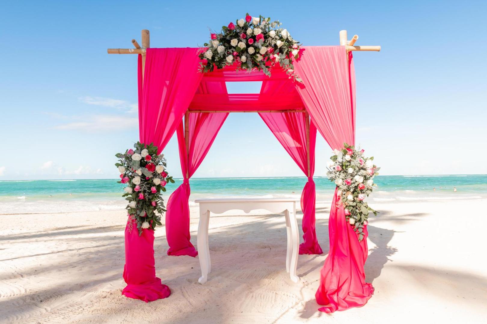 Spilgti rozā kāzu nojume ar ziedu dekorācijām baltu smilšu pludmalē.