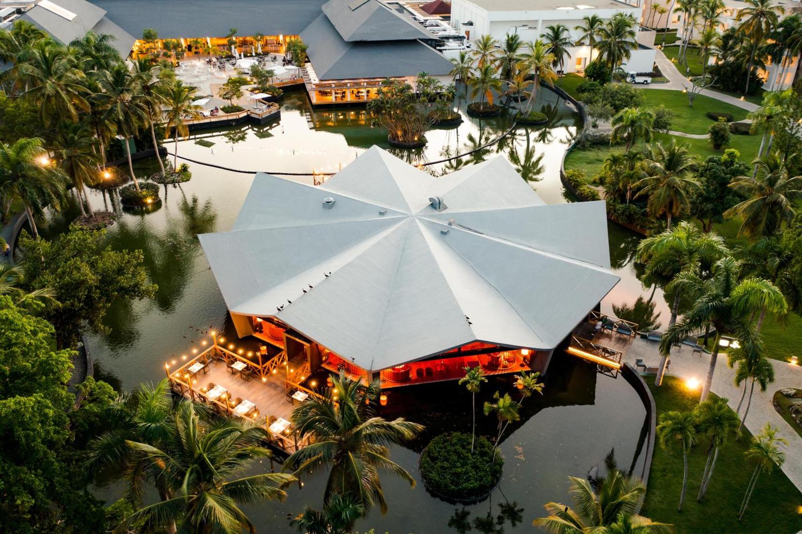 Aerofoto skats uz modernu viesnīcas restorānu, kas atrodas dīķa vidū starp palmām.
