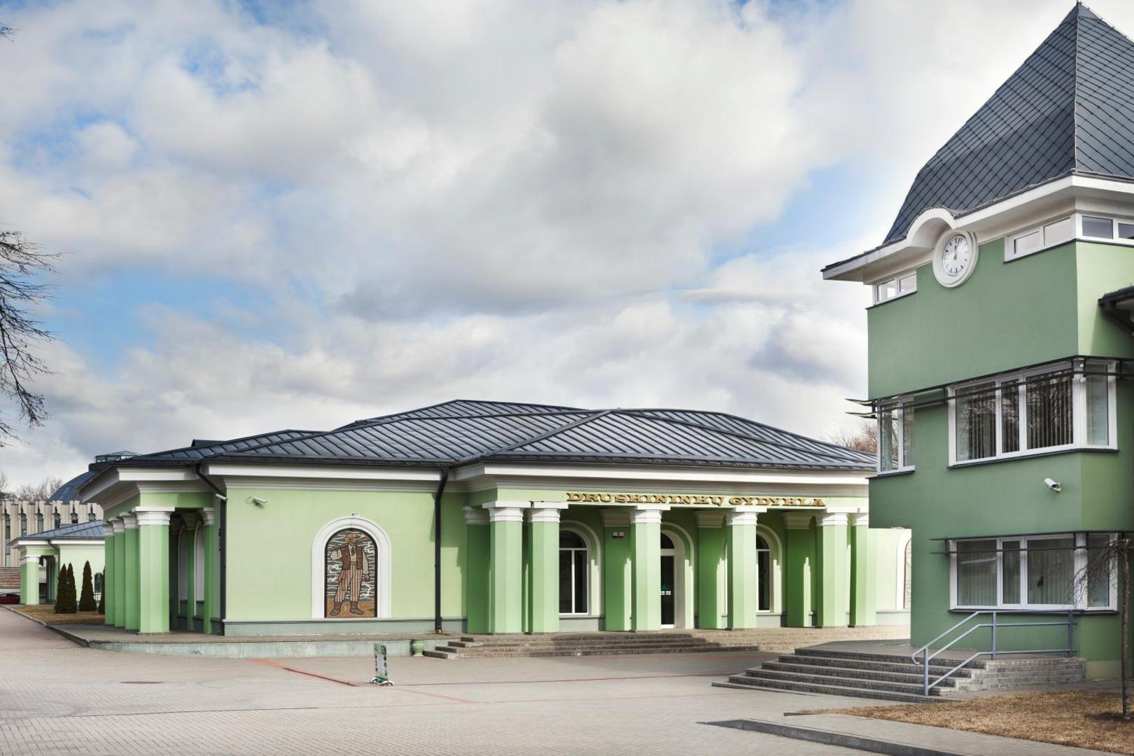 Viesnīcas Flores Hotel klasiskā stila ēka Druskininkos ar gaiši zaļu fasādi, kolonnām un dekoratīvu mozaīku pie ieejas.