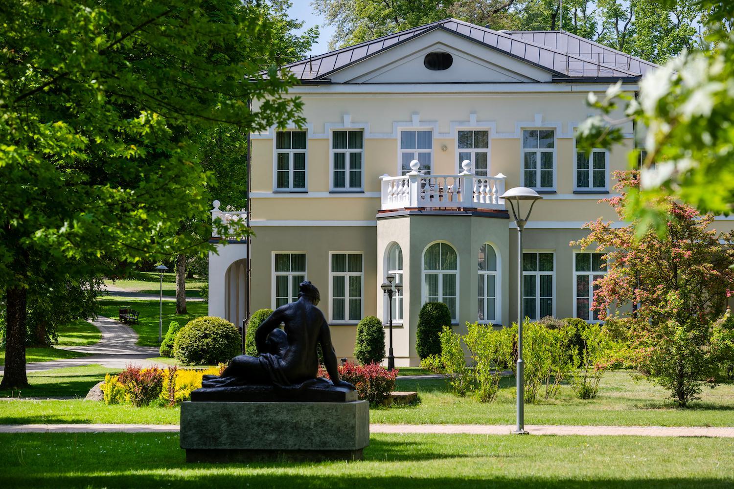 Europa Royale Druskininkai klasiskā stila korpuss ar balkonu un skulptūru priekšplānā, ko ieskauj zaļš zālājs un koki
