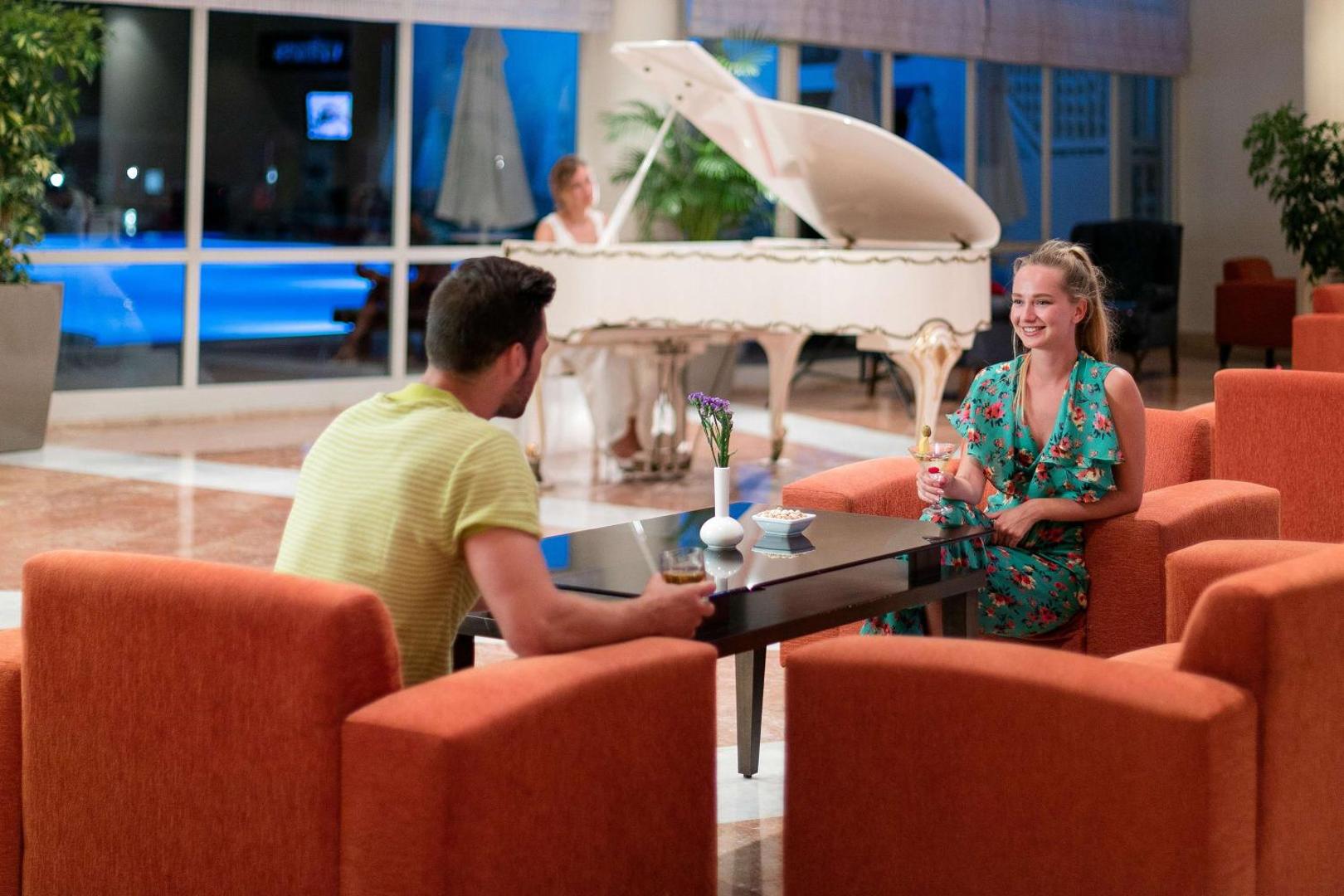 Seven Seas Hotel Life lobby bārs ar baltu flīģeli un pāri, kas bauda kokteiļus oranžos atpūtas krēslos vakarā