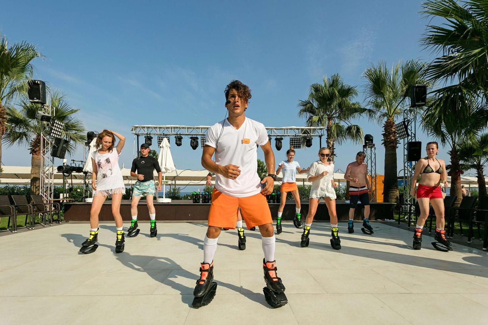 Fitnesa instruktors un viesi izpilda vingrojumus Kangoo Jumps nodarbībā uz āra skatuves Seven Seas Hotel Life teritorijā