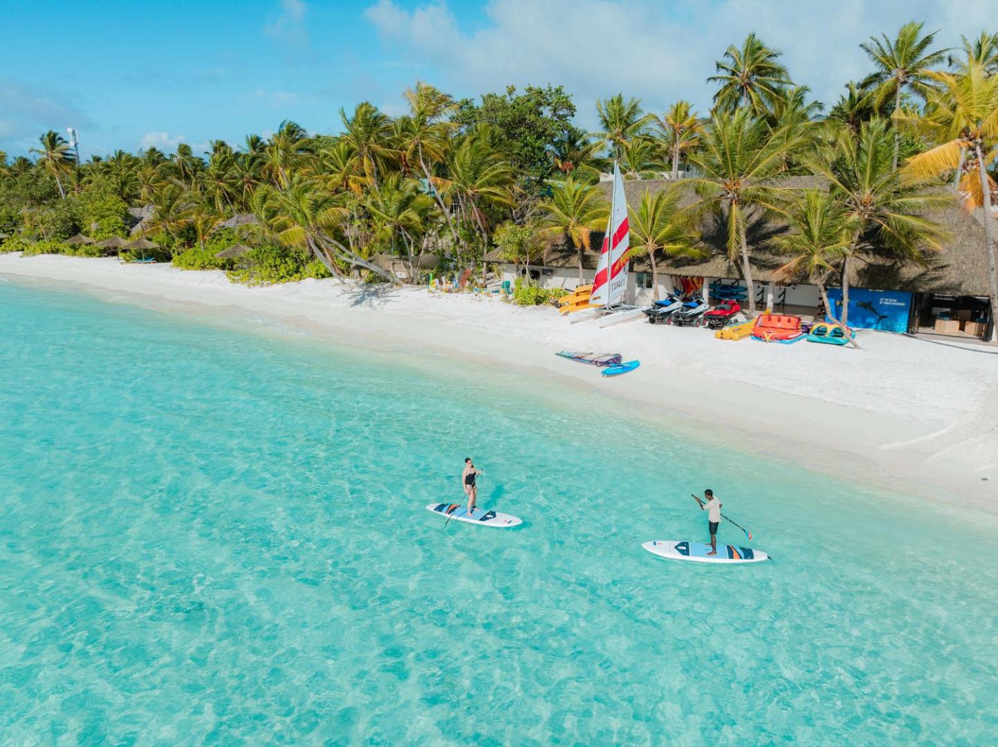 Veligandu Maldives Resort Island pludmale no putna lidojuma ar SUP dēļiem dzidrā ūdenī un ūdens sporta centru krastā