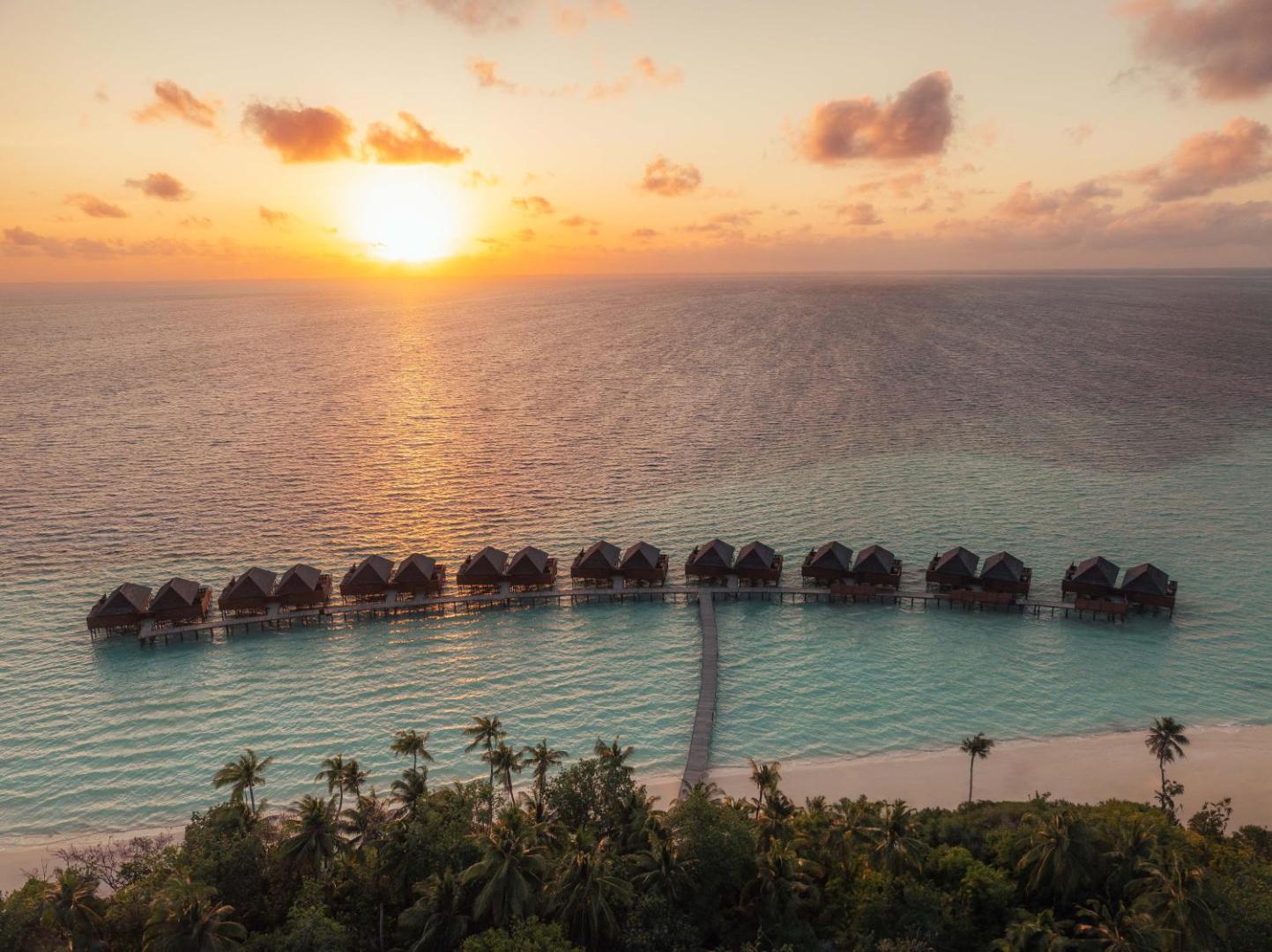 Veligandu Maldives Resort Island skats no putna lidojuma uz ūdens bungalo rindu saulrieta laikā virs tirkīza lagūnas un palmu birzs