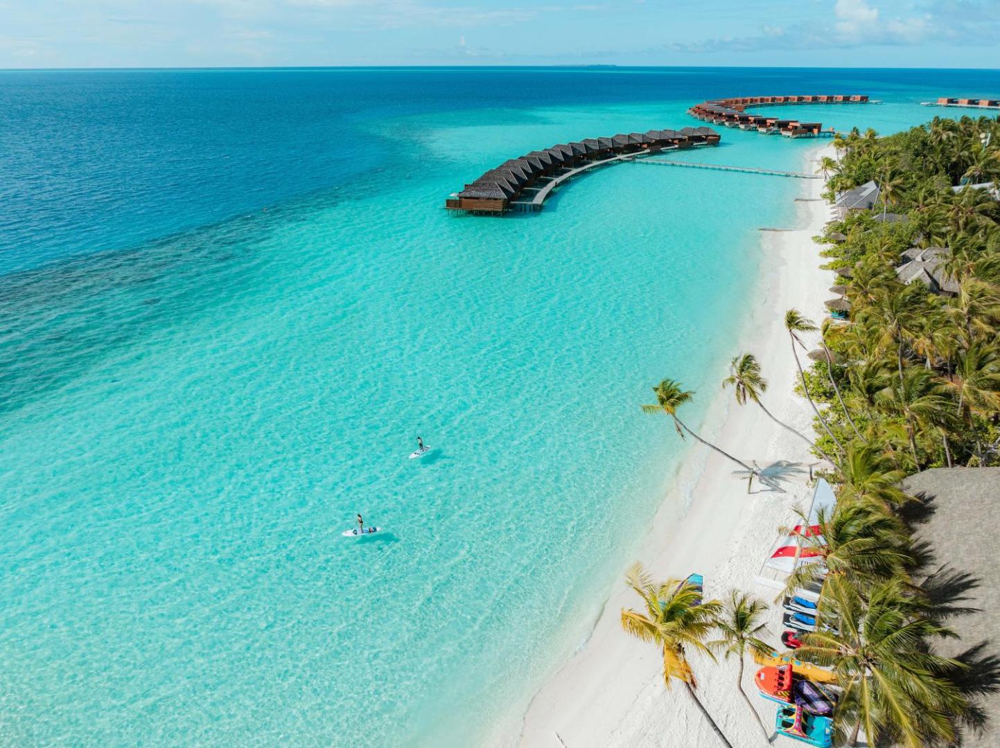 Veligandu Maldives Resort Island pludmale ar baltām smiltīm, palmām un ūdens sporta inventāru — tirkīza ūdens un bungalo tālumā