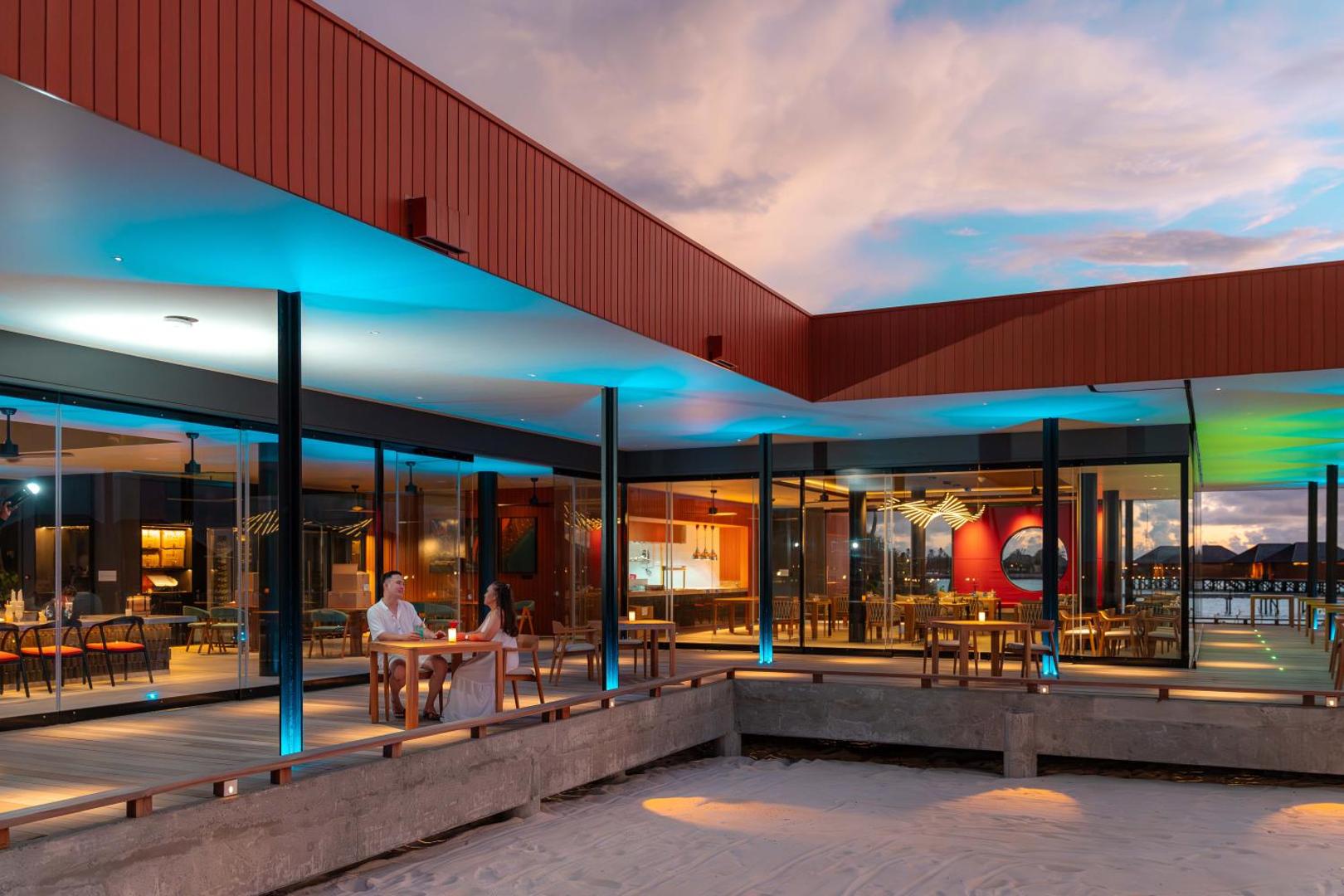 Veligandu Maldives Resort Island modernā restorāna āra terase vakarā ar stilīgu apgaismojumu un galdiņiem diviem pie smilšu pludmales