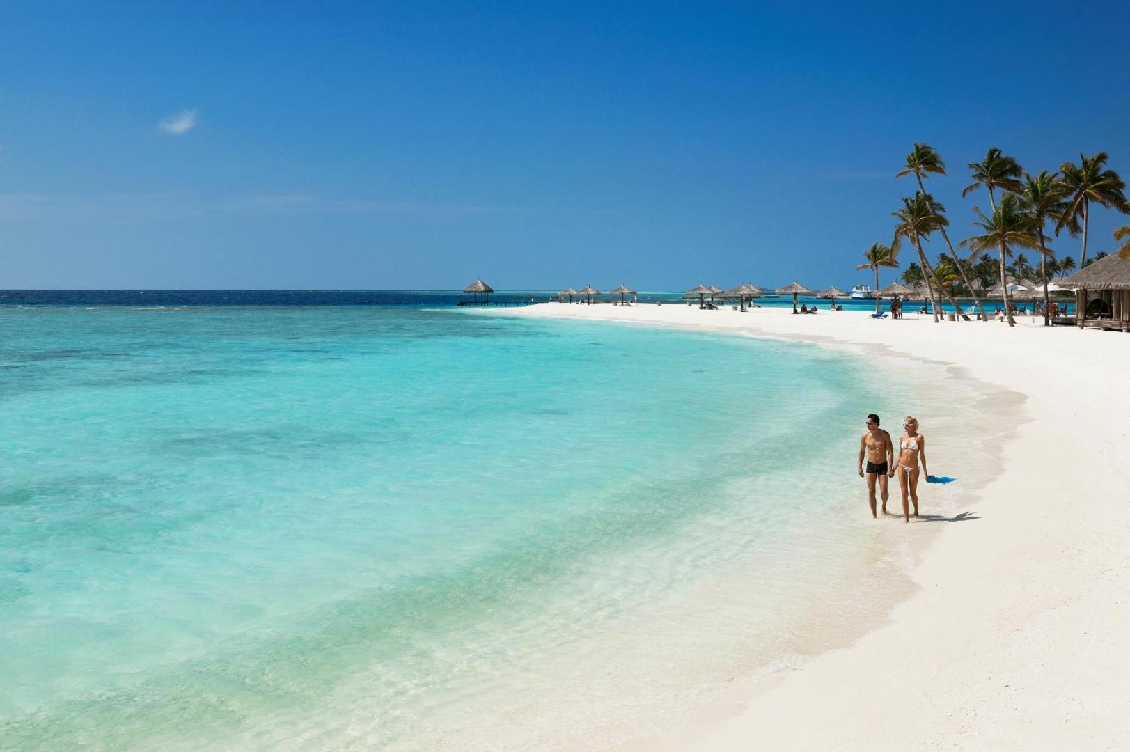 Veligandu Maldives Resort Island sniegbaltā smilšu pludmale un tirkīzzils okeāns ar pāri, kas pastaigājas gar krastu saulainā dienā