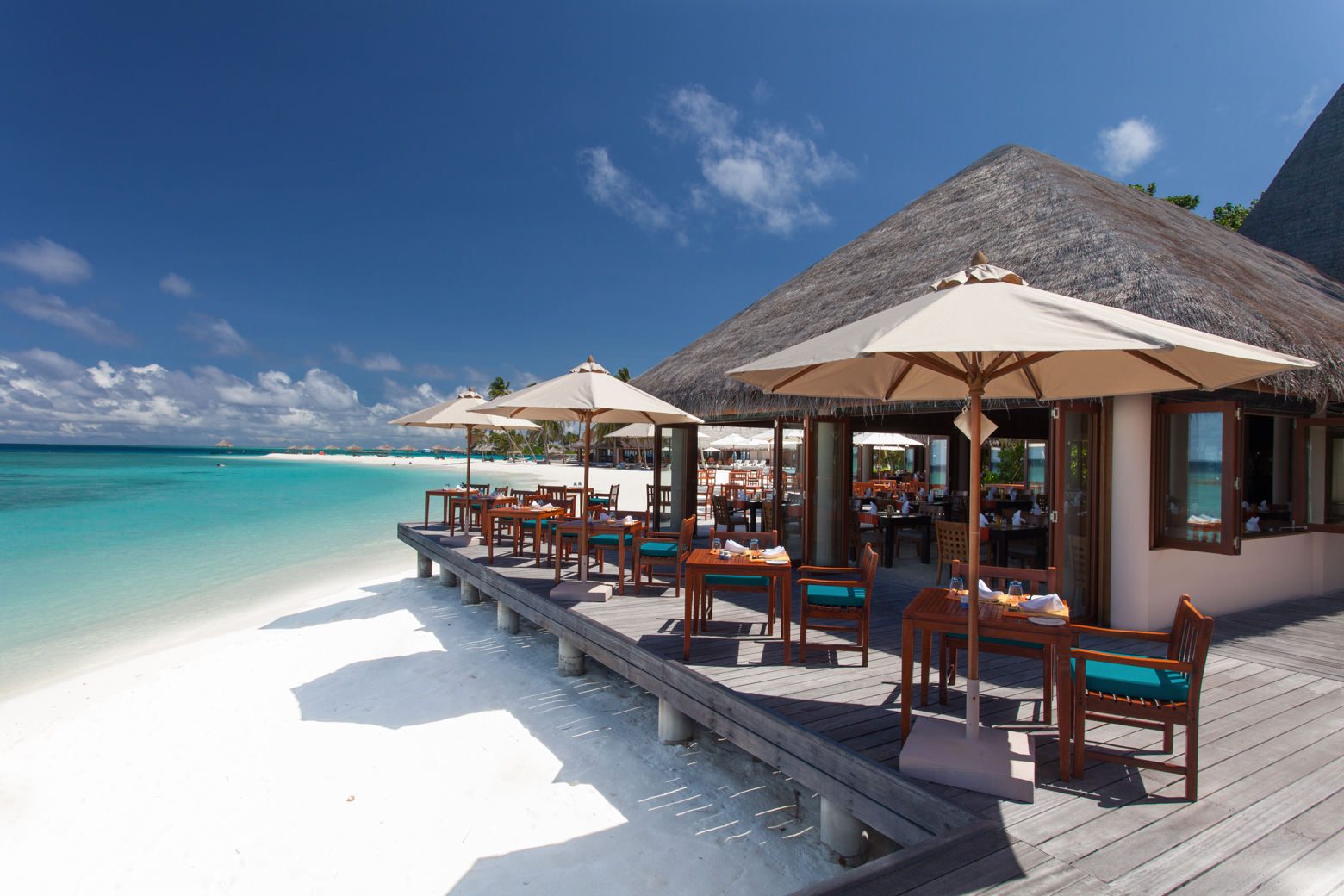 Veligandu Maldives Resort Island pludmales restorāns ar koka terasi, saulessargiem un galdiņiem tieši pie baltām smiltīm un zila okeāna