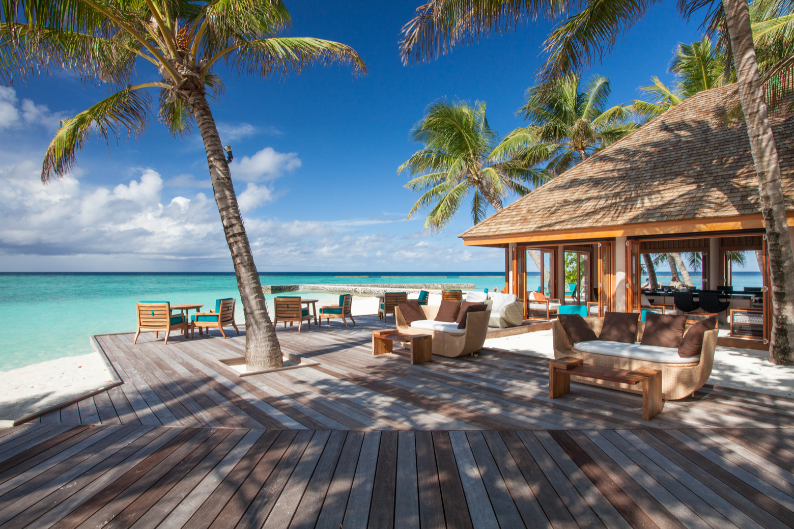 Veligandu Maldives Resort Island atpūtas zona pie okeāna ar koka terasi, ērtiem dīvāniem un palmām tirkīza ūdens piekrastē