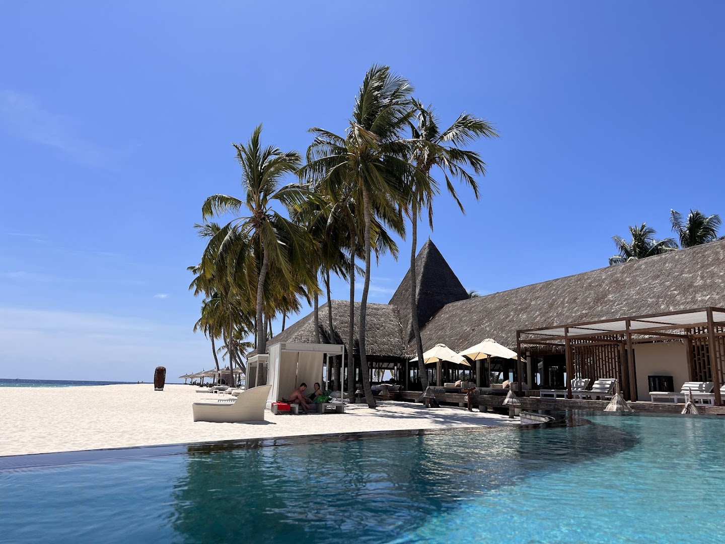 Veligandu Maldives Resort Island pludmales zona ar bezgalības baseinu, atpūtas nojumēm un tradicionāla stila ēkām pie baltām smiltīm