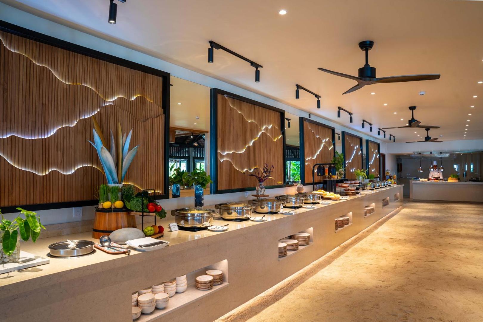 Veligandu Maldives Resort Island restorāna bufetes zona ar modernu interjeru, koka sienas paneļiem un plašu silto ēdienu izvēli metāla traukos