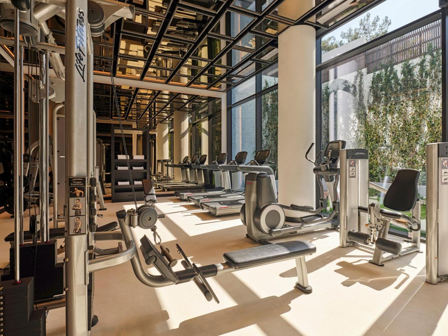 Caja by Maxx Royal modernā fitness zāle ar Life Fitness skrejceļiem un spēka trenažieriem gaišā telpā ar skatu uz dārzu.