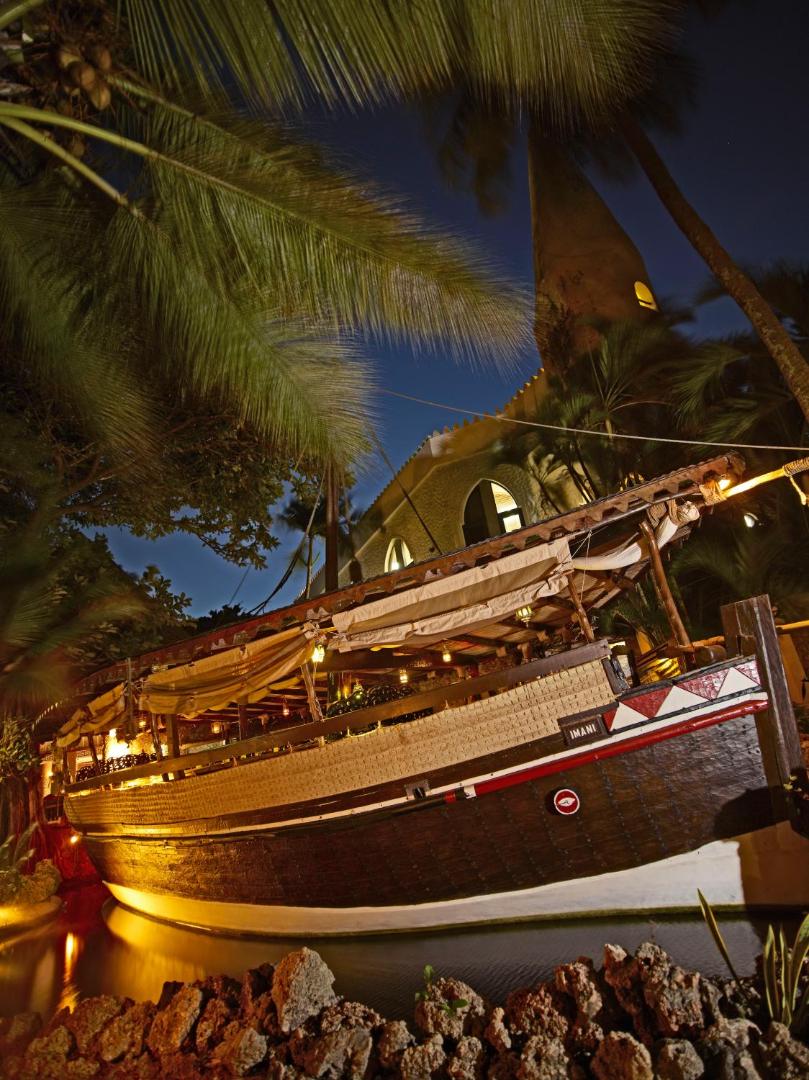 Tradicionāls dhow kuģis Imani dārzā Severin Sea Lodge viesnīcā Kenijā naktī — izgaismots restorāns zem palmām