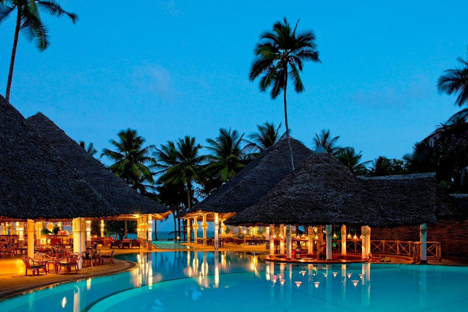 Neptune Village Beach Resort āra baseins vakarā ar izgaismotiem atpūtas namiņiem un palmām Zanzibāras piekrastē, Tanzānijā