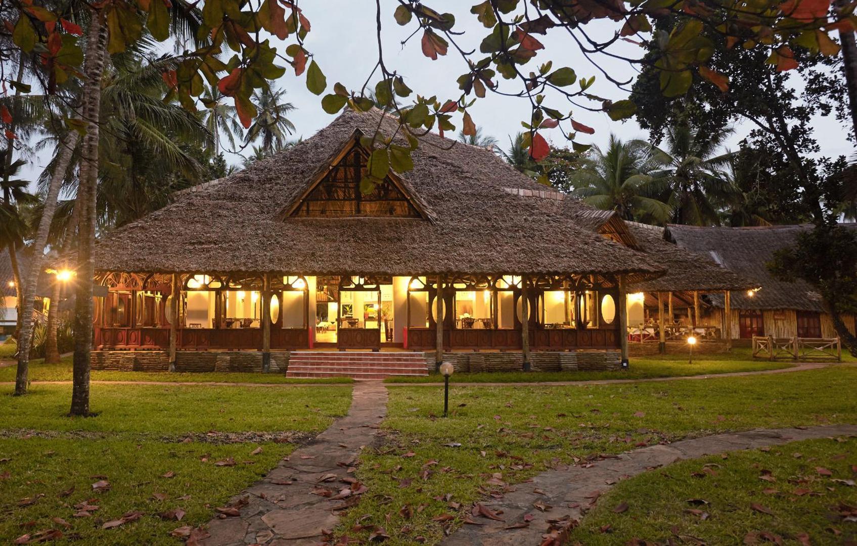 Neptune Village Beach Resort galvenā ēka ar tradicionālu makuti niedru jumtu vakarā Zanzibāras piekrastē, ieskauta palmu dārzā.