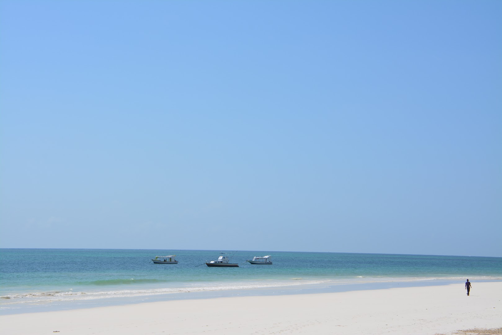 Neptune Village Beach Resort sniegbaltā smilšu pludmale ar tirkīzzilu ūdeni un nelielām laivām pie horizonta Zanzibārā