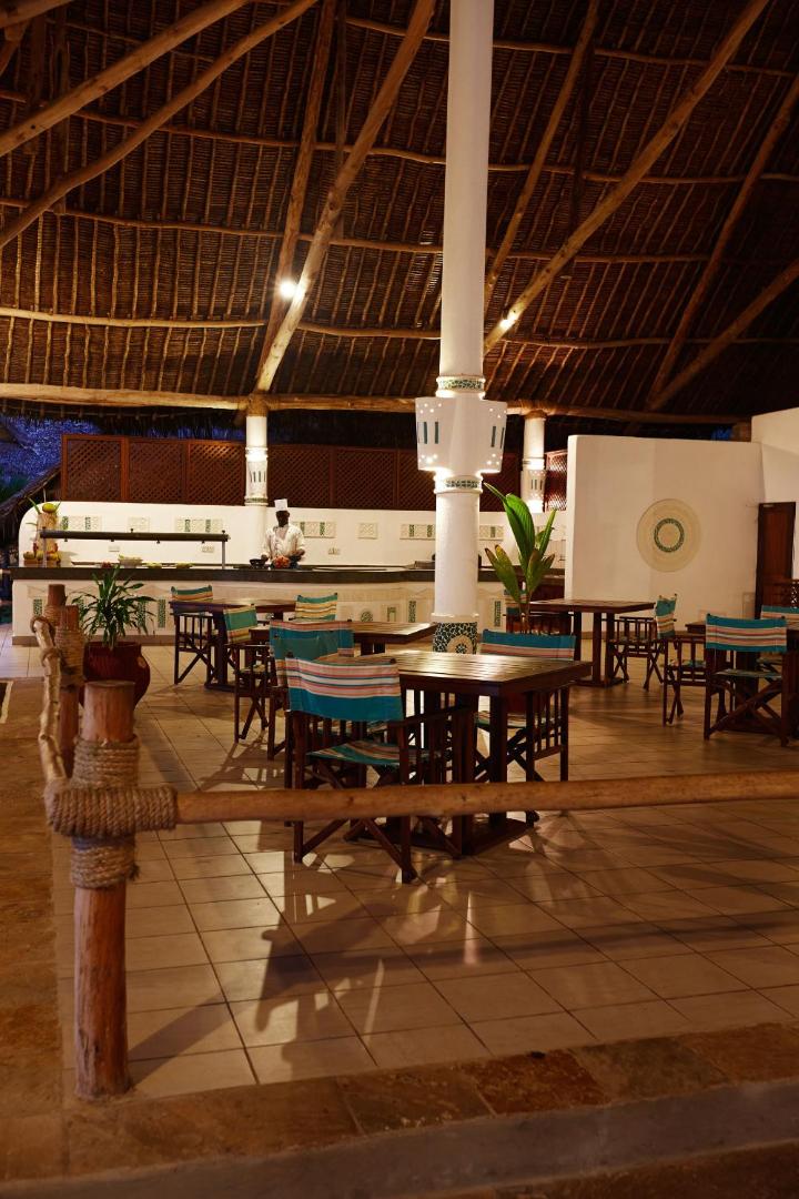 Neptune Village Beach Resort restorāna terase ar koka galdiem un krēsliem zem augstiem niedru griestiem Zanzibārā.