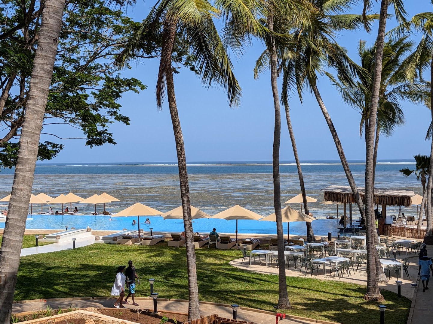 Bamburi Beach Hotel āra baseins pie okeāna krasta Mombasā, Kenijā — sauļošanās krēsli zem saulessargiem un augstas palmas dārzā.