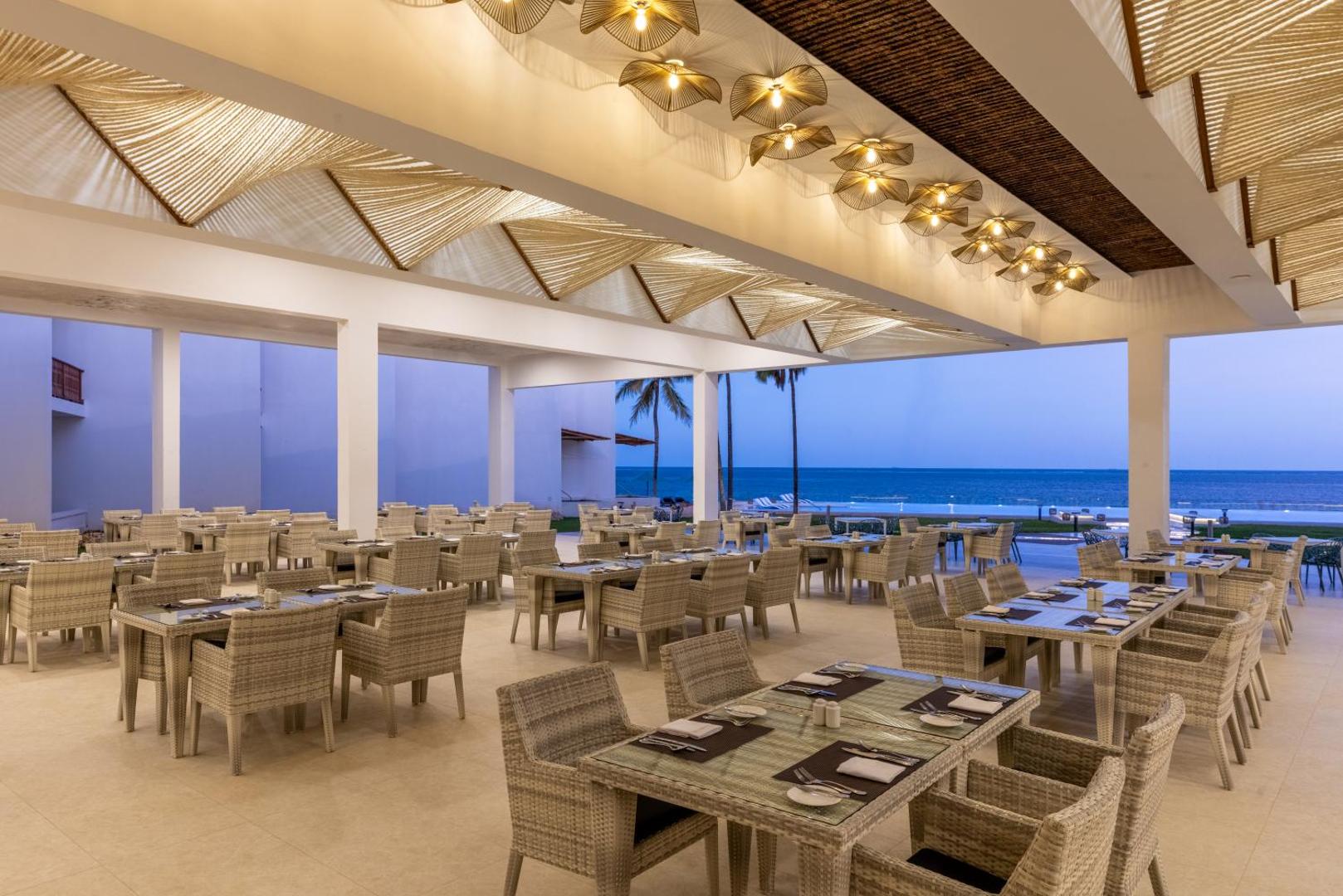 Bamburi Beach Hotel restorāns ar pītiem krēsliem un modernu apgaismojumu, no kura paveras skats uz okeānu vakarā.
