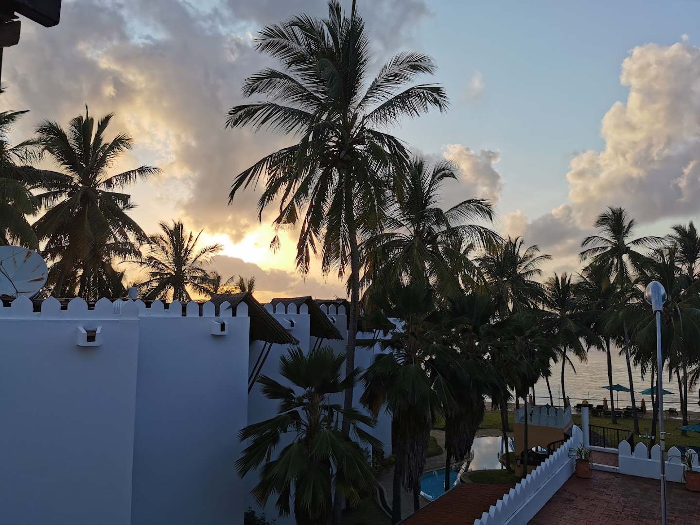 Saulriets Bamburi Beach Hotel teritorijā ar palmu siluetiem un skatu uz okeānu no viesnīcas terases vakara gaismā.