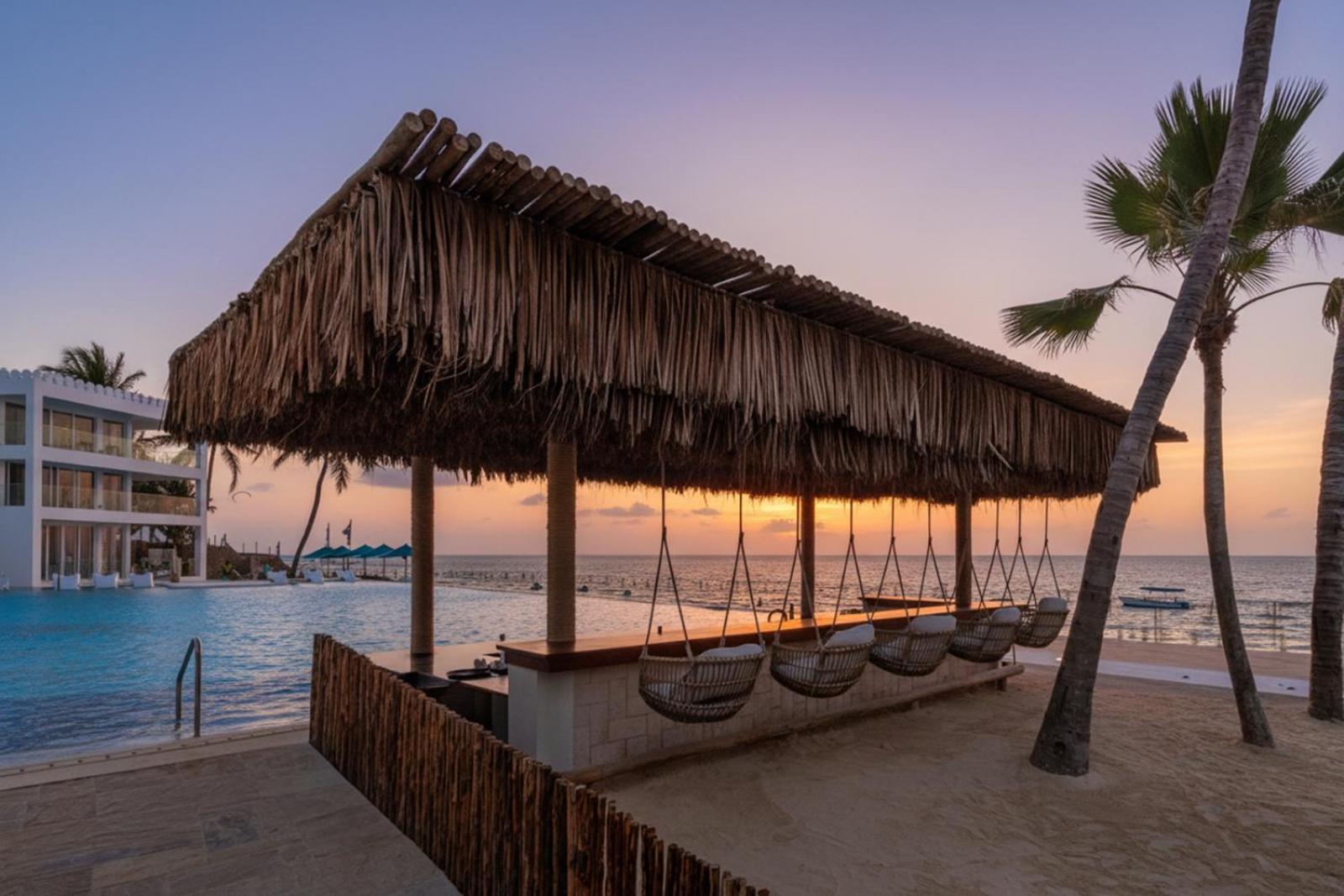 Bamburi Beach Hotel āra bārs ar salmu jumtu un šūpuļkrēsliem pie baseina saulrieta laikā Indijas okeāna krastā.
