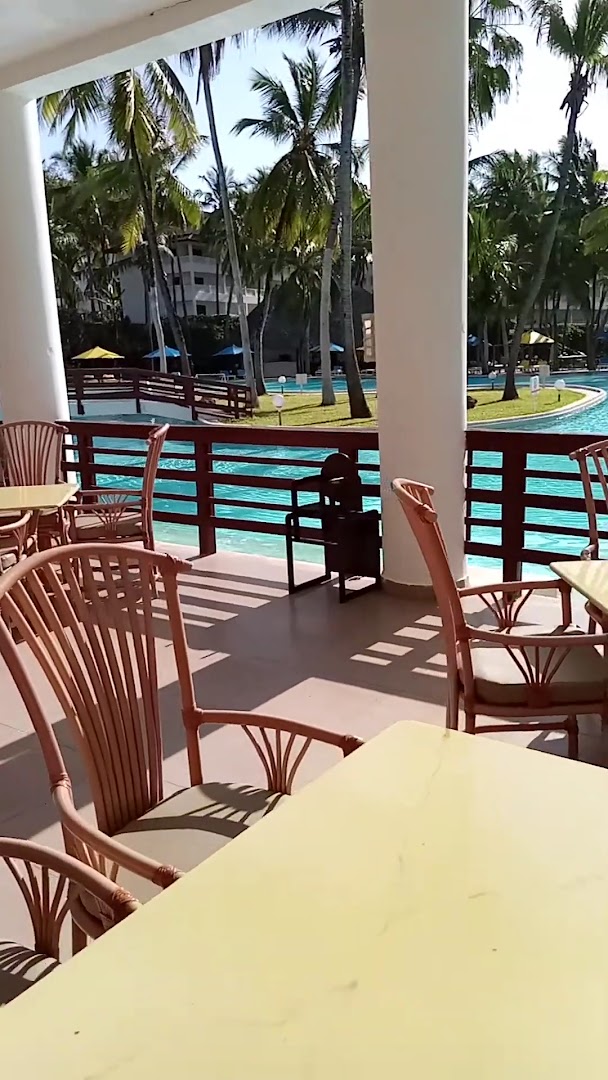 Skats no PrideInn Flamingo Beach Resort & Spa terases uz zilo baseinu un palmu dārzu saulainā dienā