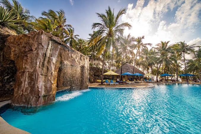 PrideInn Flamingo Beach Resort & Spa baseins ar mākslīgo ūdenskritumu un klintīm — saulrieta gaisma un palmas fonā