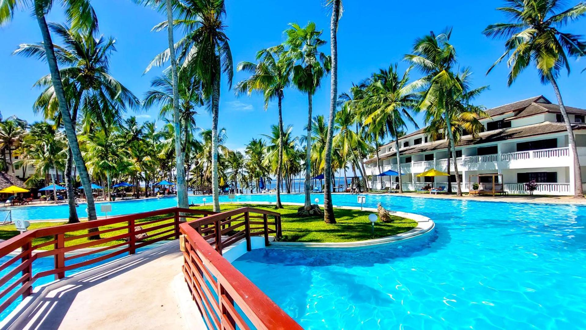 PrideInn Flamingo Beach Resort & Spa baseina zona ar koka tiltiņu un viesnīcas ēku fonā — tropu dārzs ar palmām