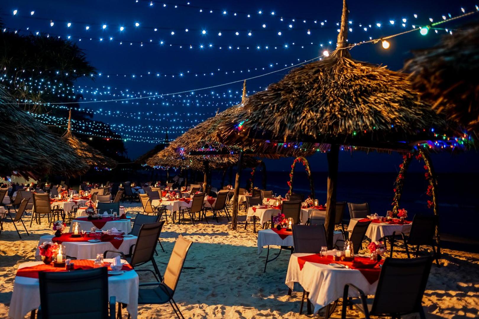 Romantiskas vakariņas Southern Palms Beach Resort pludmalē zem zvaigžņotām debesīm ar svētku apgaismojumu un klātiem galdiem smiltīs.