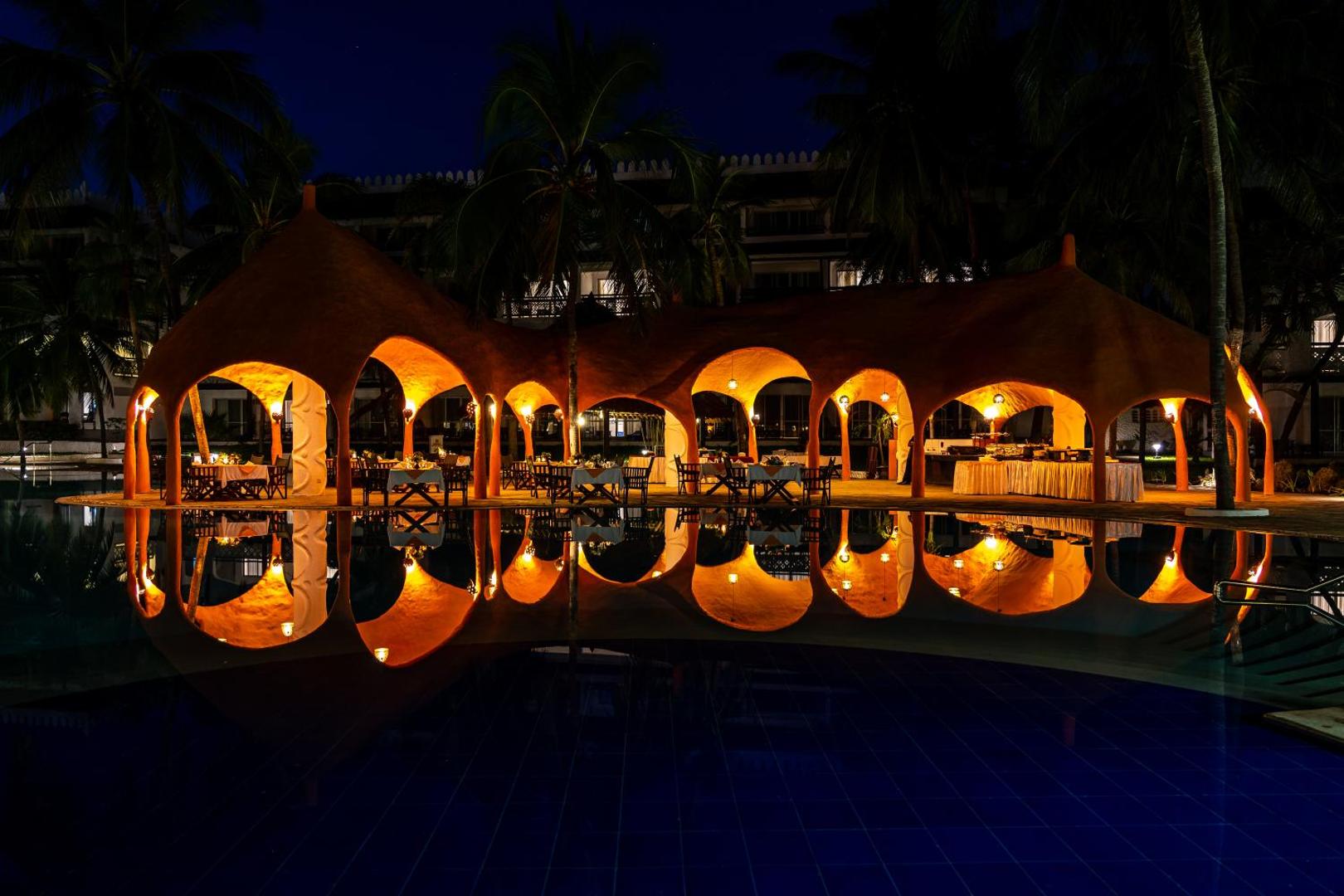 Southern Palms Beach Resort baseina zona naktī ar izgaismotu arhitektūru, arkām un palmu atspulgu mierīgā ūdens virsmā