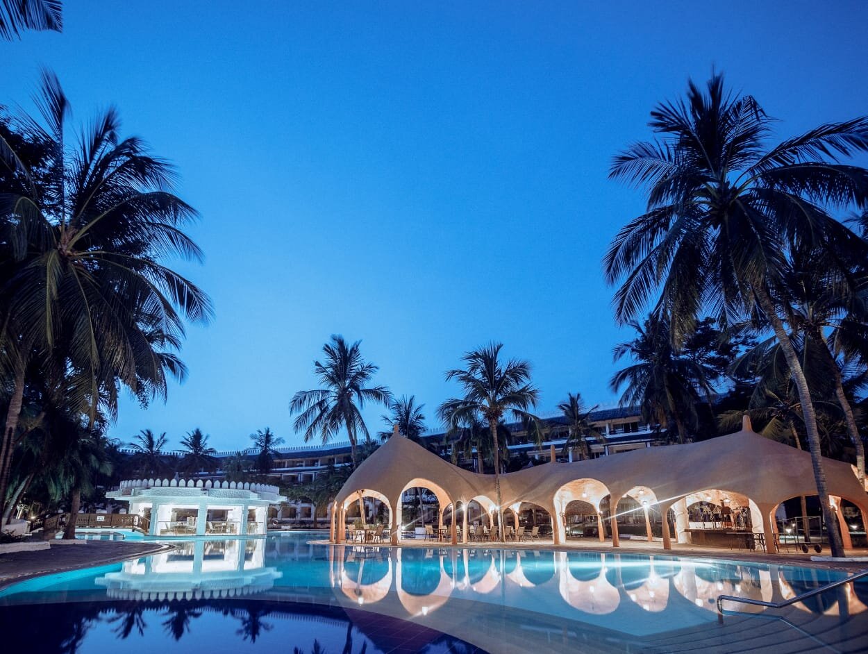 Southern Palms Beach Resort baseins un restorāna ēka vakara apgaismojumā ar atspulgiem ūdenī un augstām palmām pret zilo debesu fonu.
