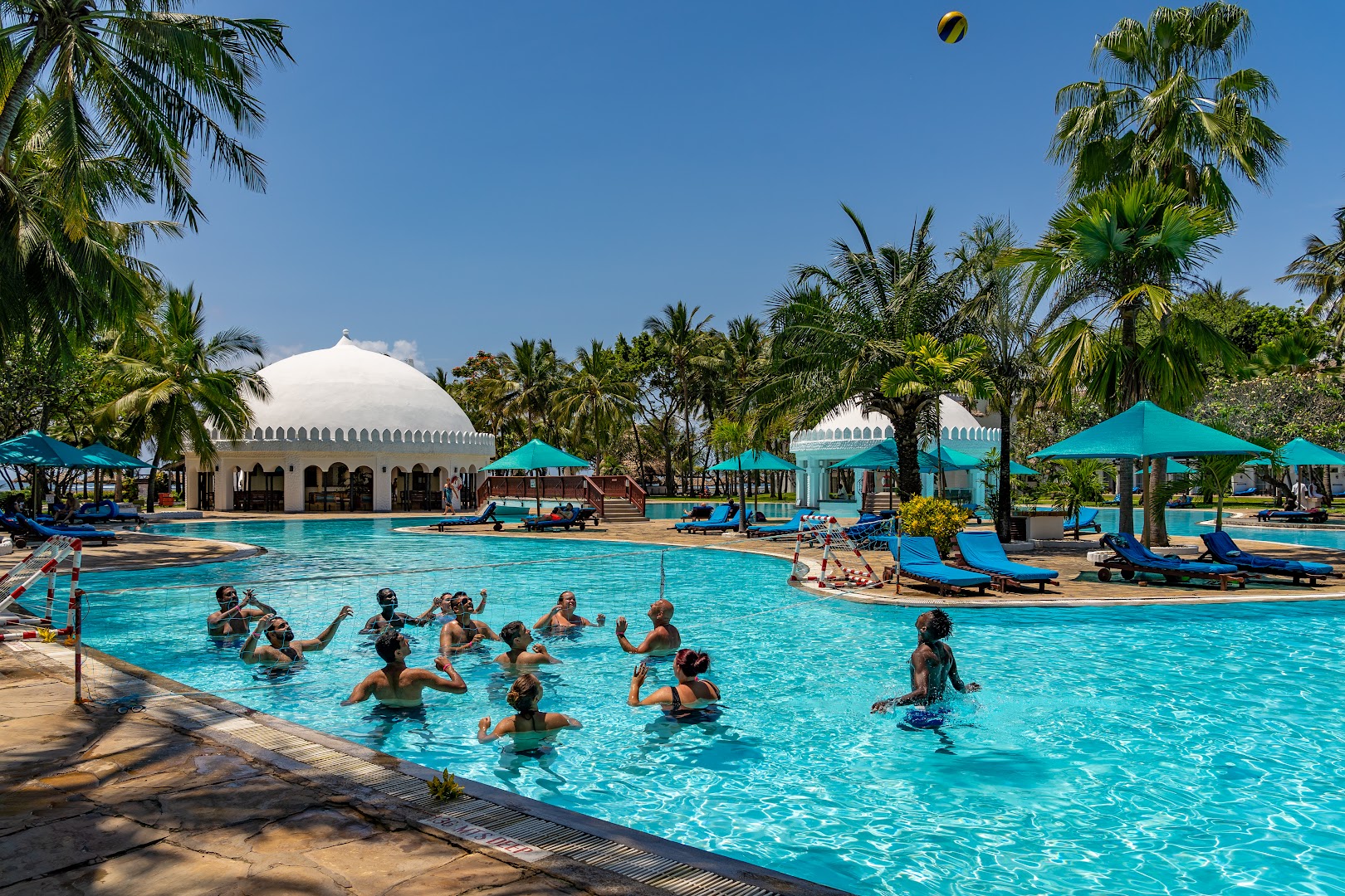 Southern Palms Beach Resort lielais āra baseins ar atpūtniekiem, kuri spēlē ūdens volejbolu, fonā palmas un balti kupolveida restorāni Kenijā