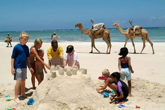 Bērni būvē smilšu pili Southern Palms Beach Resort pludmalē, fonā redzami kamieļi un tirkīzzils okeāna ūdens