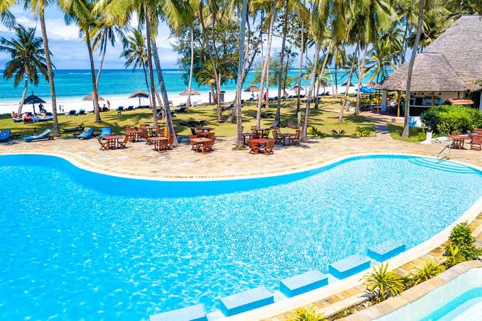 Diani Sea Lodge viesnīcas baseina zona un restorāna galdiņi zem palmām pie Indijas okeāna krasta Kenijā