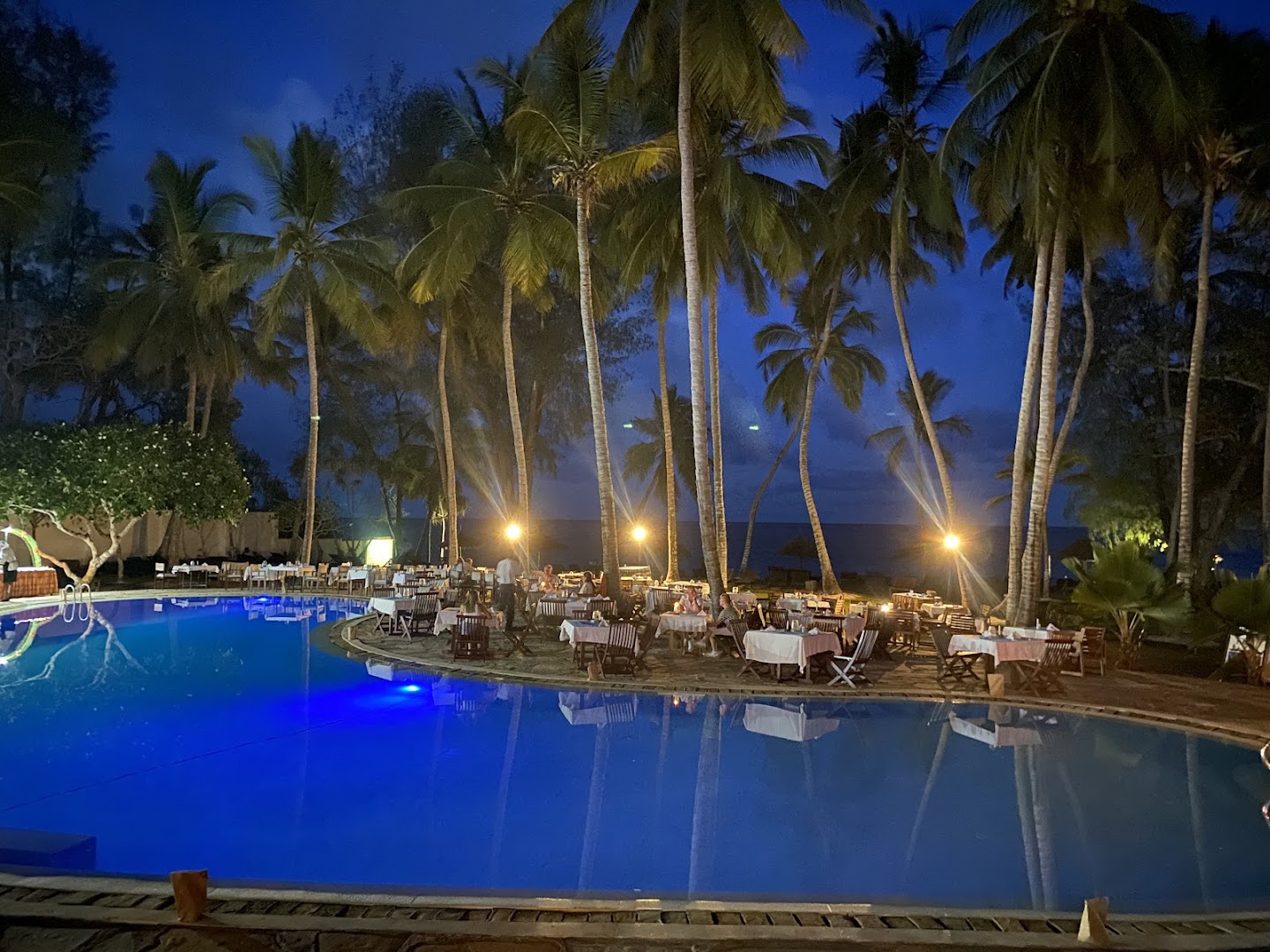 Diani Sea Lodge āra baseins un restorāna zona vakarā ar izgaismotām palmām un galdiņiem piekrastē, Kenijā