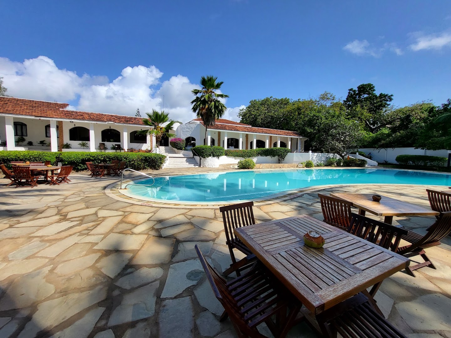 Diani Sea Lodge viesnīcas ēka ar terasi un koka galdiņiem pie baseina saulainā dienā Kenijā